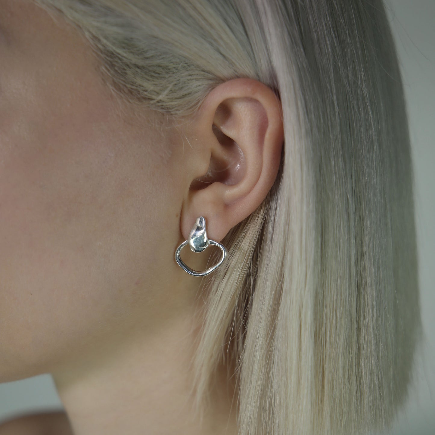 The Circle Drops earrings are a handmade creation crafted from 925 sterling silver. The irregular, smooth, and glossy circle at the bottom creates a unique formation, while the wide silver strip at the top adds a subtle, smooth, and shiny element.
