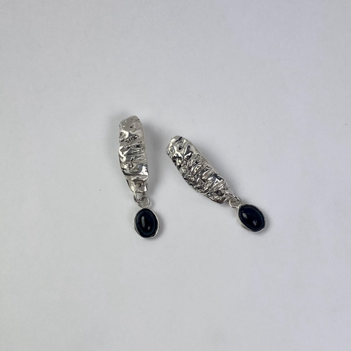 The Malva earrings are handmade and crafted from sterling silver 925, showcasing a raw texture and form that highlight their natural beauty. Their glossy finish adds an extra touch of radiance. The earrings are joined with a setting accompanied by a semi-precious stone known as black agate.