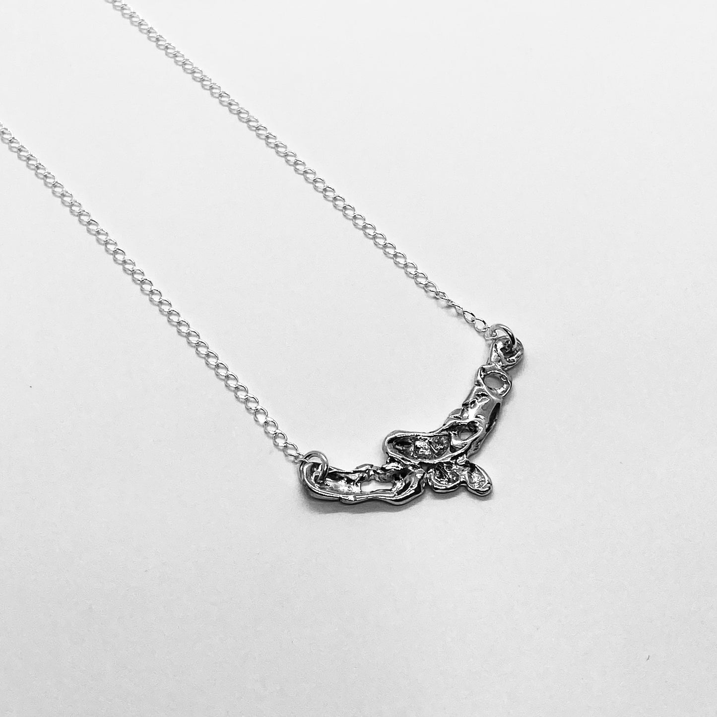 The Camelie necklace is a handmade piece crafted from sterling silver 925. Its design is minimalistic with a raw and glossy appearance, featuring various openings in its interior. Accompanied by a delicate silver chain, the length in the photo is 42 centimeters, and you have the option to choose your preferred length.