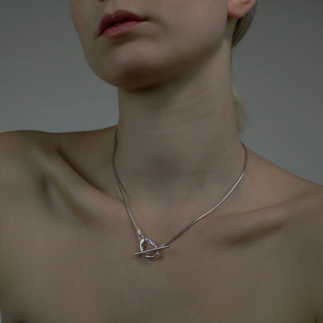 The Kyoto necklace is a handmade piece crafted from 925 sterling silver. Its chain is thick and connected to a small, untreated circle. The fastening is located on the front side