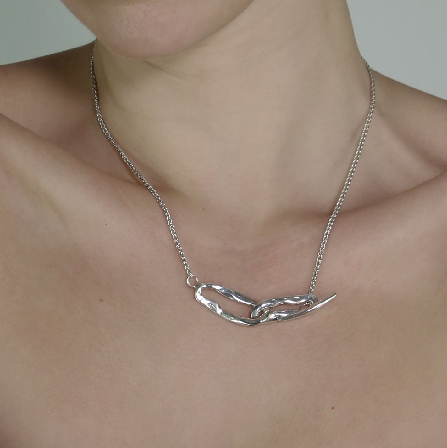 The Calma necklace is a handmade piece crafted from sterling silver 925. It features two irregular silver shapes connected by a thin rod that intricately weaves through them, creating a unique and secure closure. The texture resembles raw elegance and has a glossy finish.
