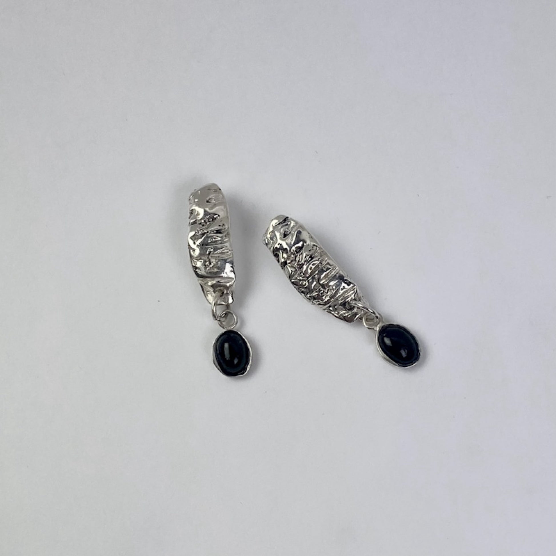 The Malva earrings are handmade and crafted from sterling silver 925, showcasing a raw texture and form that highlight their natural beauty. Their glossy finish adds an extra touch of radiance. The earrings are joined with a setting accompanied by a semi-precious stone known as black agate.