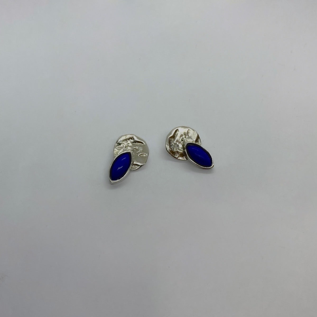 The Violet earrings is handmade and made of 925 sterling silver. Its base is circular with a raw texture, accompanied by an embedded blue semi-precious stone in the shape of a drop