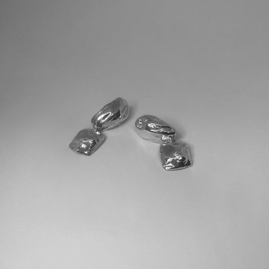 The Follaje earrings are handmade and crafted from sterling silver 925. They have a natural, untreated texture and consist of two parts. The upper part has an oval shape and is connected to a square, irregularly shaped element that is joined with a small hoop. They are polished for a smooth finish.
