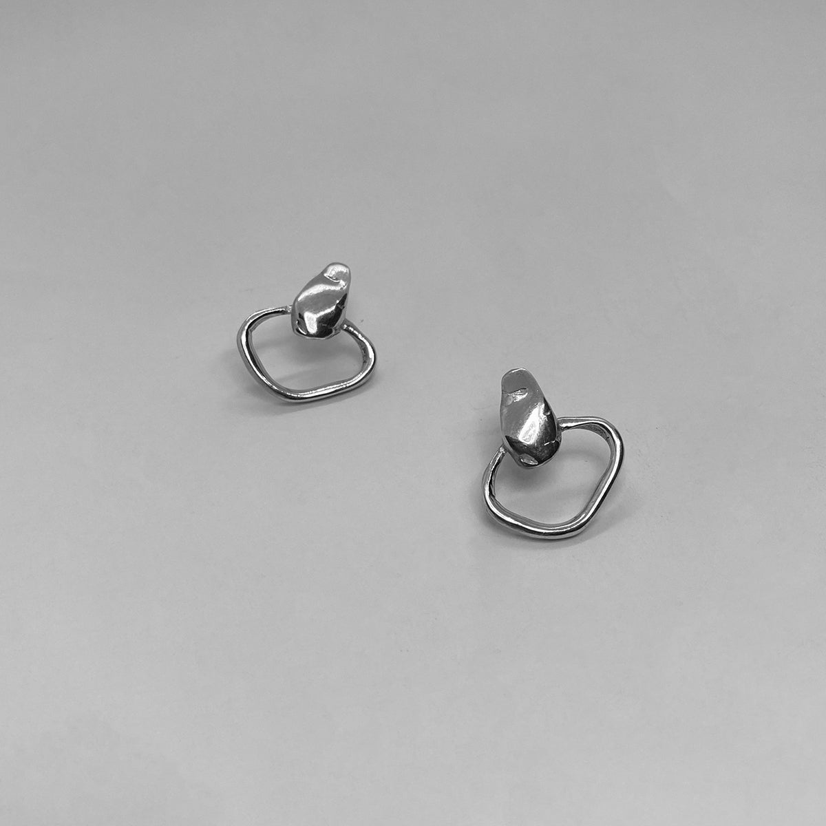 The Circle Drops earrings are a handmade creation crafted from 925 sterling silver. The irregular, smooth, and glossy circle at the bottom creates a unique formation, while the wide silver strip at the top adds a subtle, smooth, and shiny element.